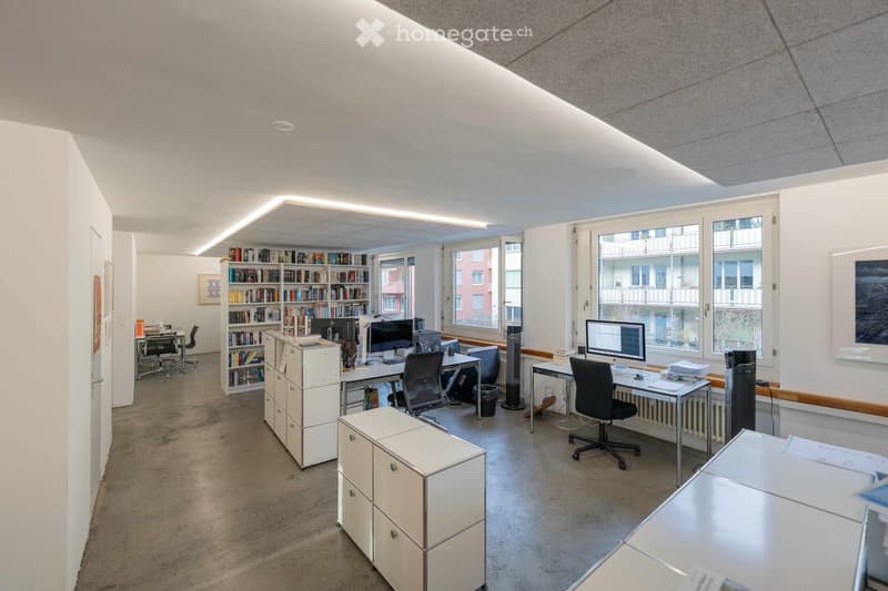Modern office with large windows, bookshelves, office desks, computer monitors, chairs, radiators