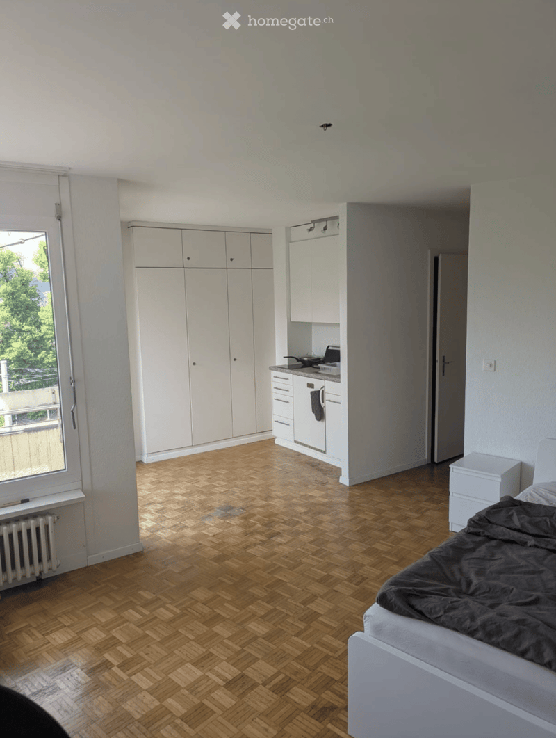 Bright bedroom with a wooden herringbone floor, a white bed with dark bedding, built-in white kitchen cabinets, a small stove, and a radiator under the window.
