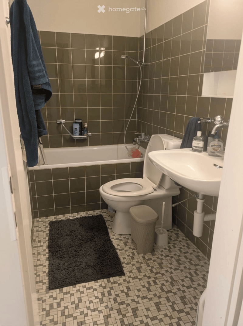 Bathroom with dark green tiled walls, white fixtures, and a black bath mat on the floor.
