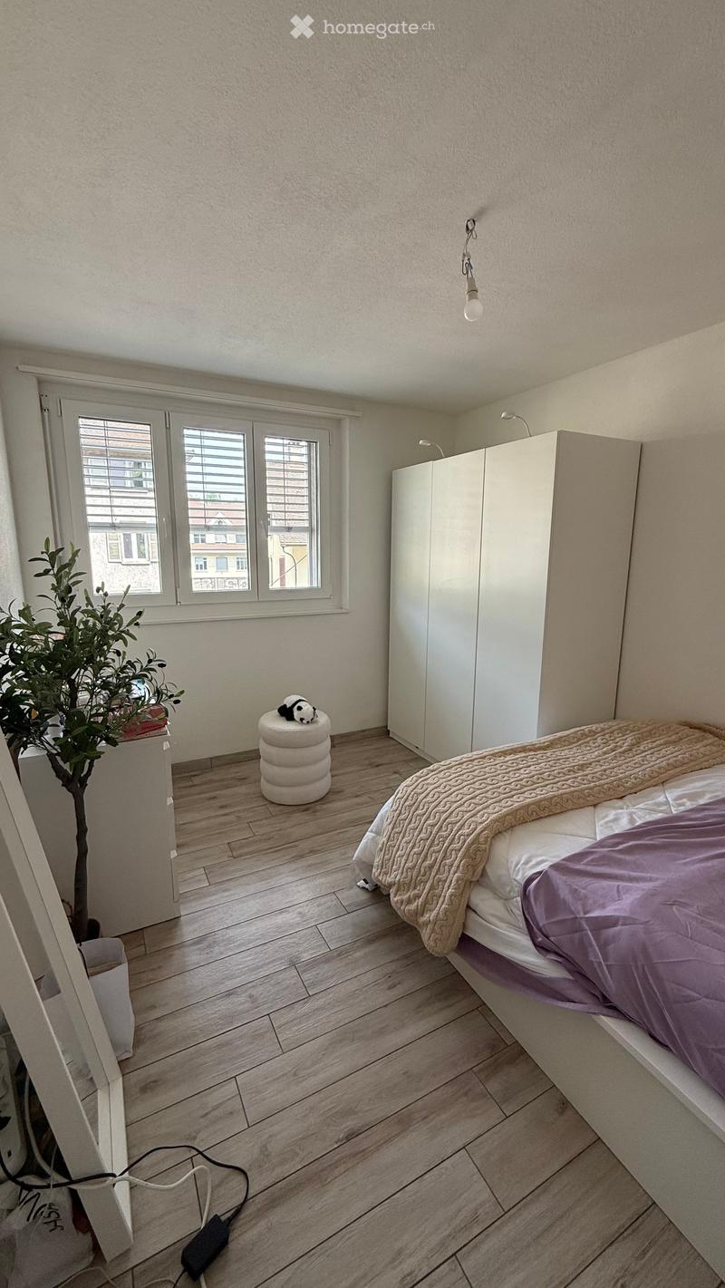 Cozy bedroom with wooden flooring, a bed with a purple blanket, a white wardrobe, a small stool with a panda toy, and a potted plant near the window.