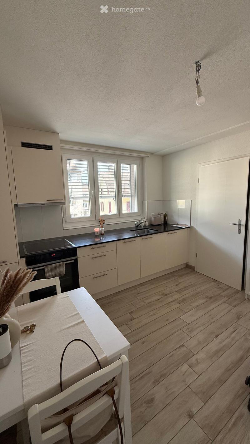 Modern kitchen with light-colored cabinets, a black countertop, and a white dining table with chairs. The floor is wooden, and there is a window with blinds.
