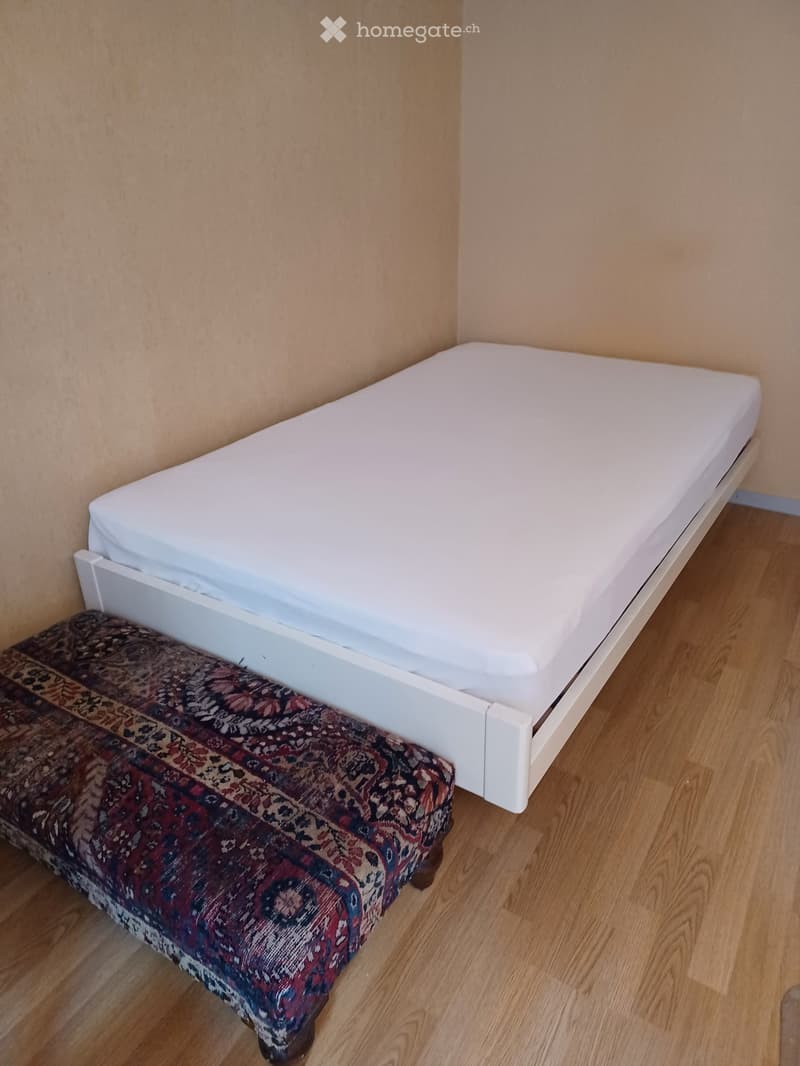 A small, furnished room with a single bed and a patterned bench at the foot of the bed. The room has light wooden flooring and beige walls.