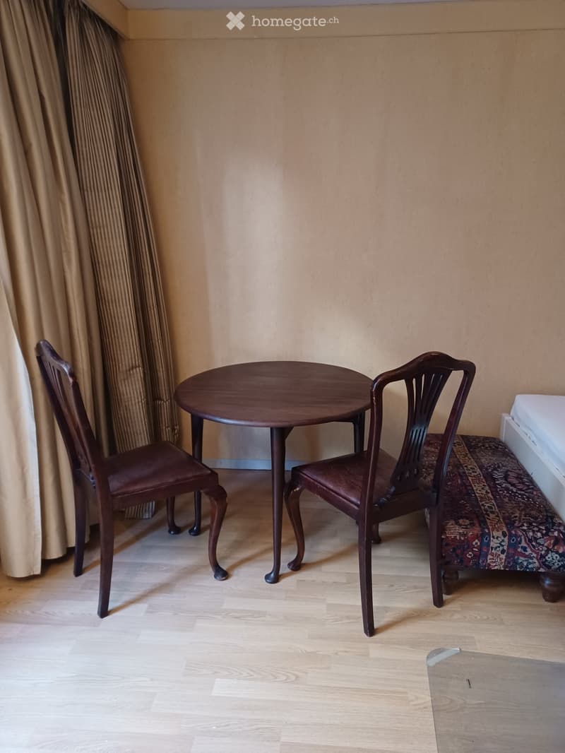 A small, furnished room with a round wooden table and two matching chairs, light wooden flooring, and beige walls.