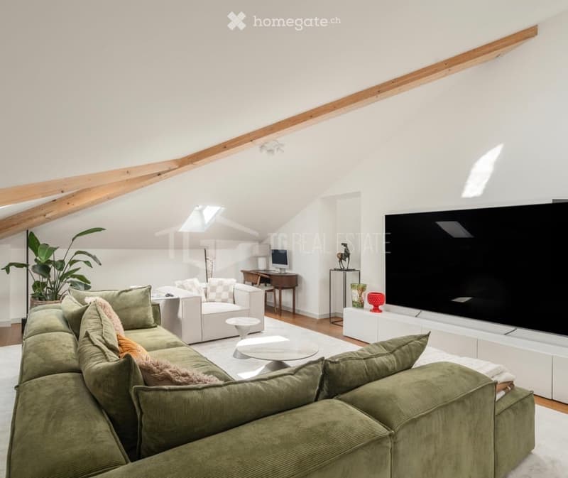 Modern living room with a large sectional sofa, coffee table, armchair, TV, and a small desk area with a computer. The room features exposed wooden beams and large windows, creating a bright and airy atmosphere.