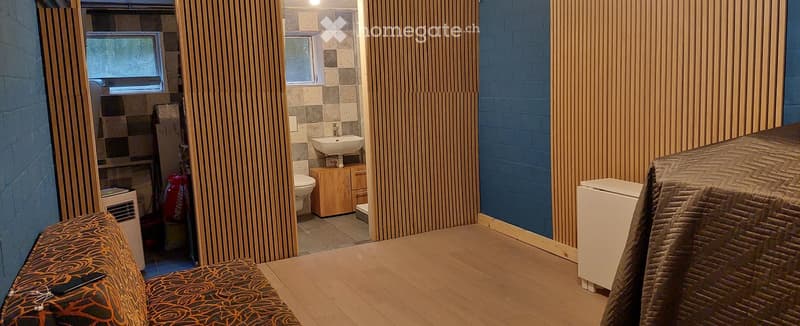 A small, furnished room with a sofa, a small table, a refrigerator, and an adjacent bathroom area with a sink, toilet, and wooden cabinet.