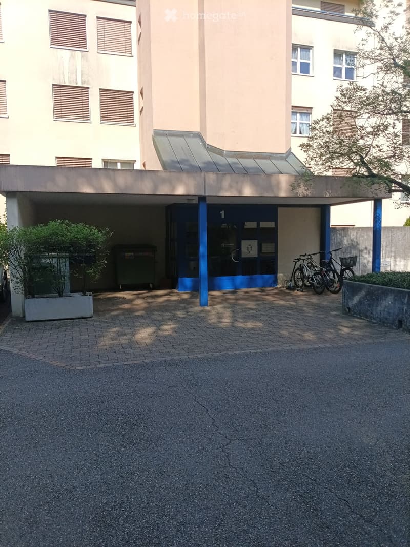 Multi-story building with a covered entrance area, bicycles parked nearby, and a small landscaped area with a tree and planter.