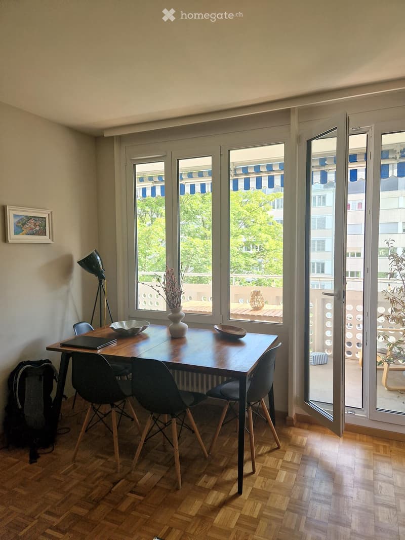 Dining area with a wooden table, four chairs, a vase with dried flowers, a lamp, and a backpack on the floor. Large windows open to a balcony with outdoor furniture.