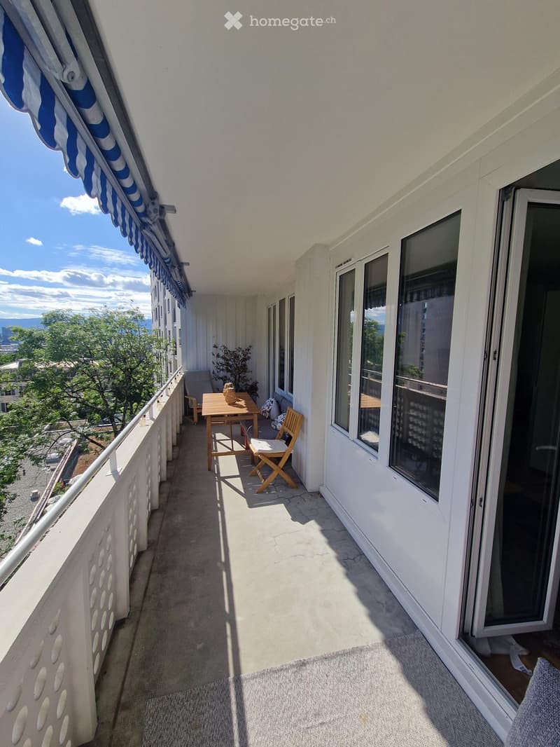 Balcony with a table, chairs, and a small stool, overlooking a cityscape with trees and buildings.