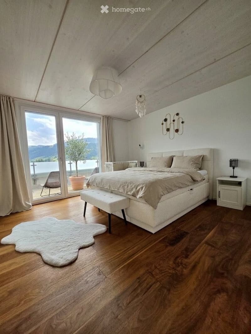 Modern bedroom with wooden flooring, large bed, nightstand, bench, and a view of a lake through large windows.