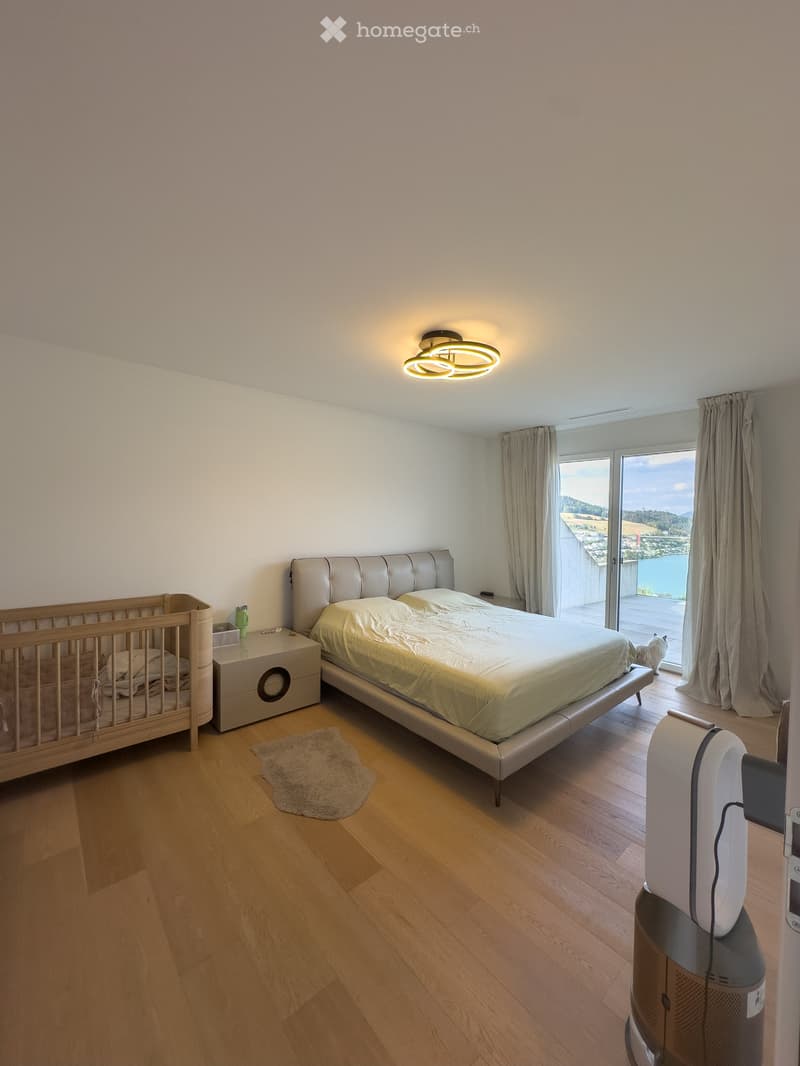 Bright bedroom with wooden flooring, a large bed, a nightstand, a wooden cradle, a fan, and a view of the lake through large windows.