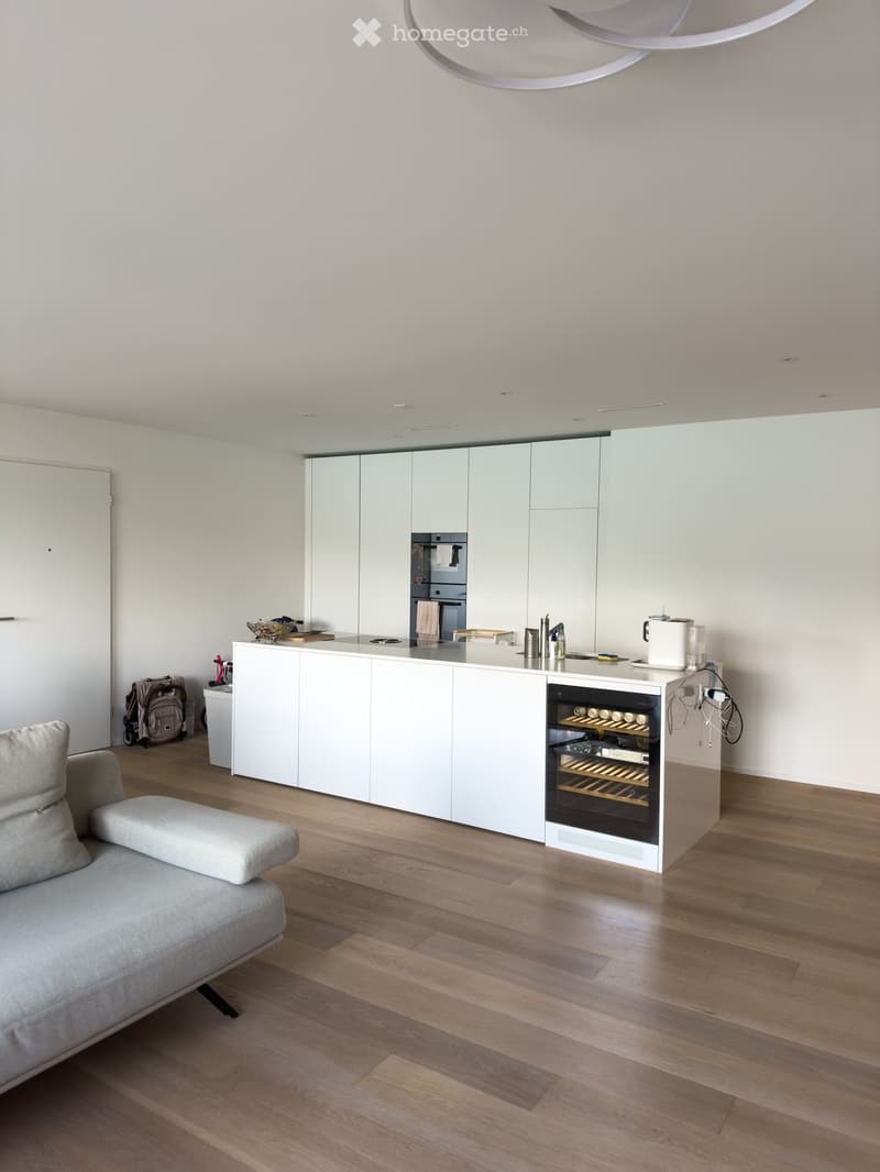 Modern, minimalist kitchen and living area with white cabinets, a central island, a wine cooler, and a grey sofa.