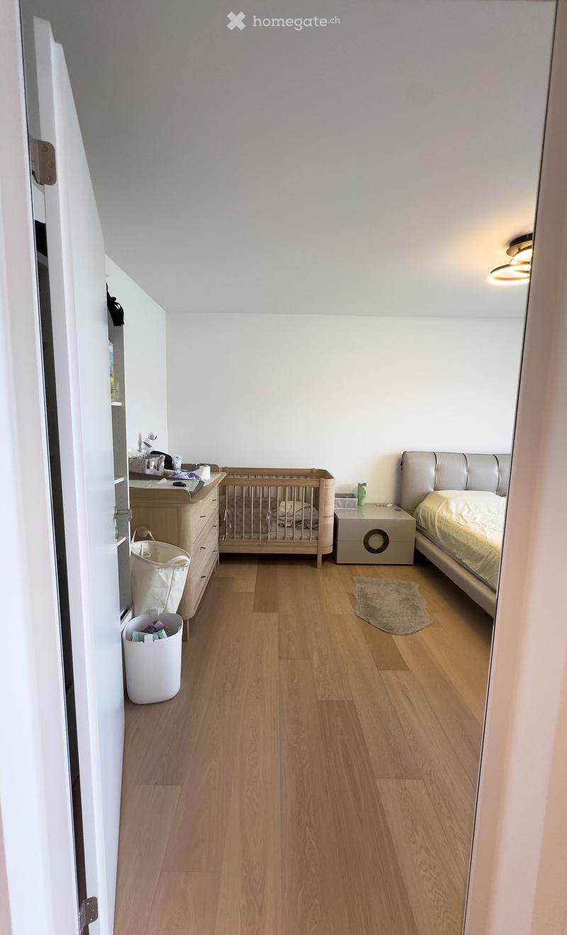 Cozy bedroom with wooden flooring, a large bed with a tufted headboard, a crib, a dresser, and a small nightstand.