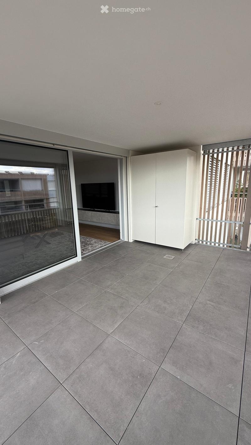 Modern balcony with grey tiled floor, white cabinet, sliding glass door leading to interior, and metal railing.