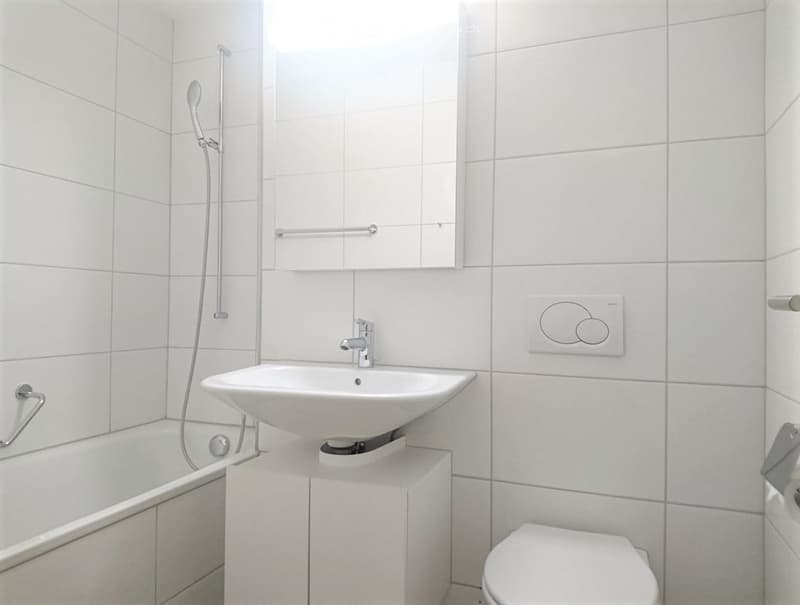 Modern bathroom with white tiled walls and floor, featuring a sink, bathtub, toilet, mirror, and showerhead.