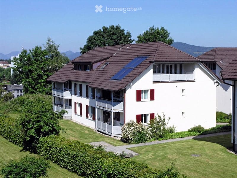 A modern two-story house with white walls, red shutters, and a brown tiled roof. Features include multiple balconies, a solar panel on the roof, and a well-maintained lawn with bushes and trees.