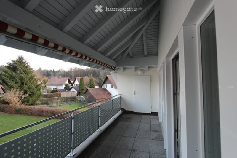 Balcony with metal railing, tiled floor, white door leading inside, view of a residential area with houses and trees.