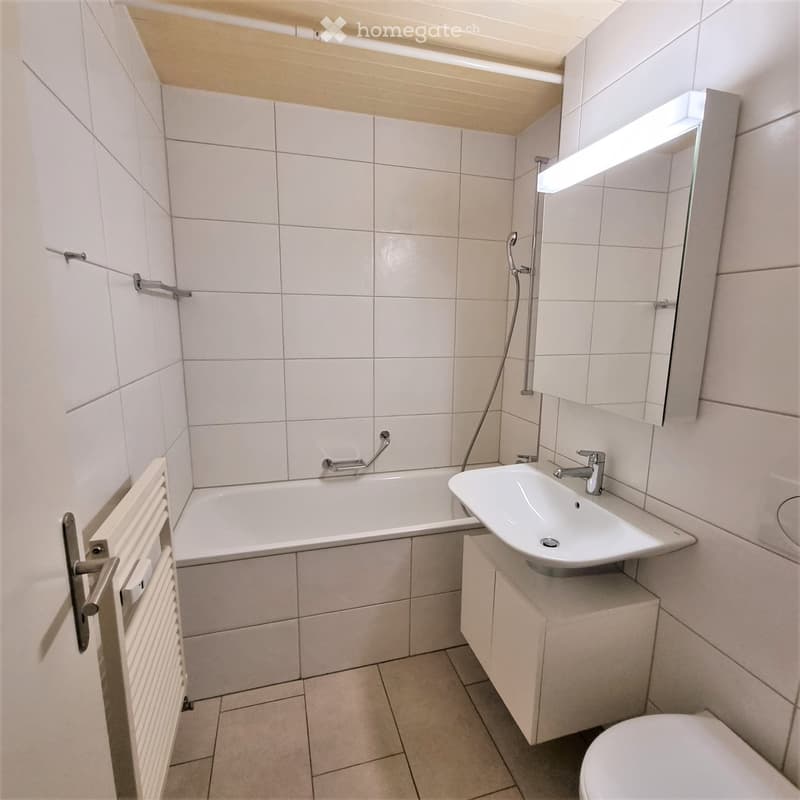 Modern bathroom with white tiles, a bathtub, a sink with a modern faucet, a toilet, a mirror with built-in lighting, and a radiator.
