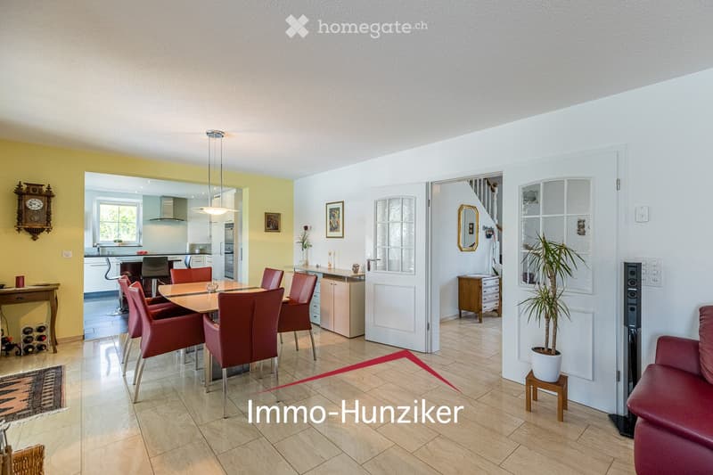 Open-plan living and dining area with a red leather sofa, a wooden dining table with red chairs, a modern kitchen area, and a wine rack. The space features light-colored walls, tiled flooring, and decorative elements like a clock and framed pictures.