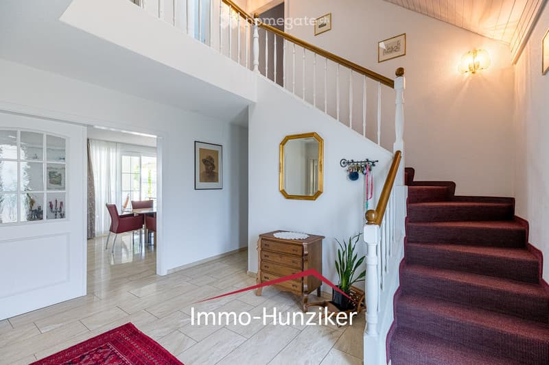 Spacious hallway with a staircase leading to an upper level, wooden railing, red carpeted stairs, a small wooden cabinet, a mirror, and a potted plant. The hallway features white walls, a tiled floor, and several framed pictures.