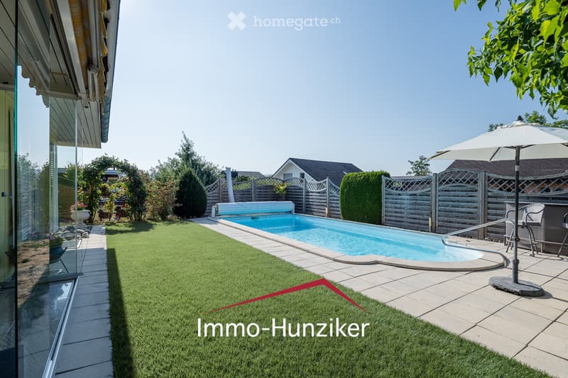 Well-maintained garden with a swimming pool, surrounded by a wooden fence, lawn, patio, and outdoor furniture including an umbrella and chairs.