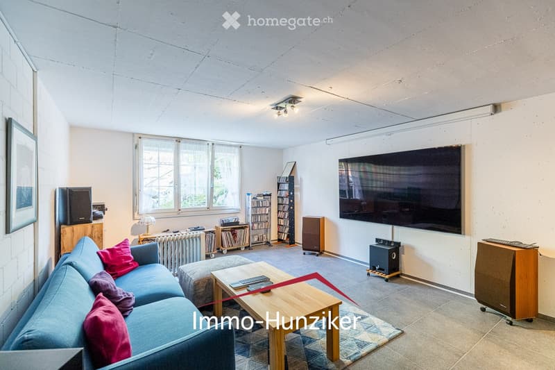 Modern living room with a blue sectional sofa, wooden coffee table, bookshelf, large screen, and speakers.