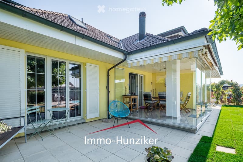 A modern glass-enclosed patio area attached to a house, featuring a dining set with chairs and a table, surrounded by a well-maintained garden with grass and potted plants.
