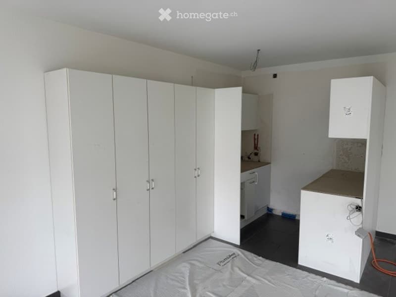 Empty room with white built-in wardrobes, a small kitchenette area with a sink and countertop, and a covered floor area, likely for a bed.