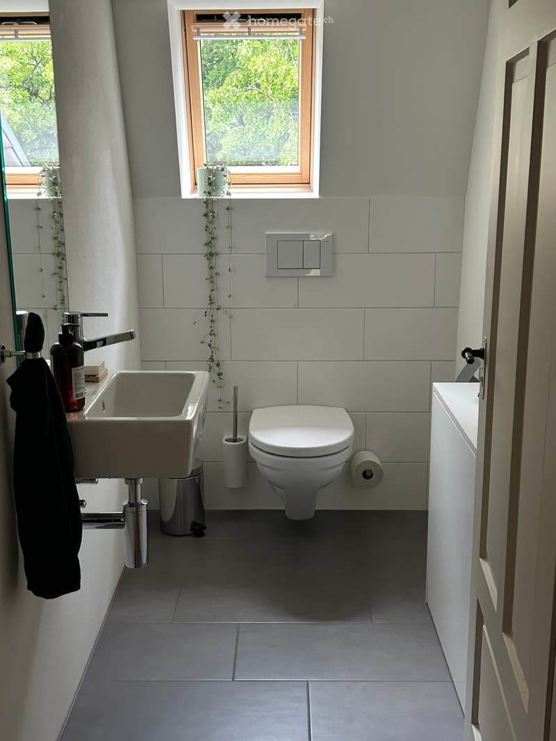 Modern bathroom with a white sink, chrome faucet, toilet, and a towel hanging on the wall. The room features light grey tiles and a window with a view of greenery.