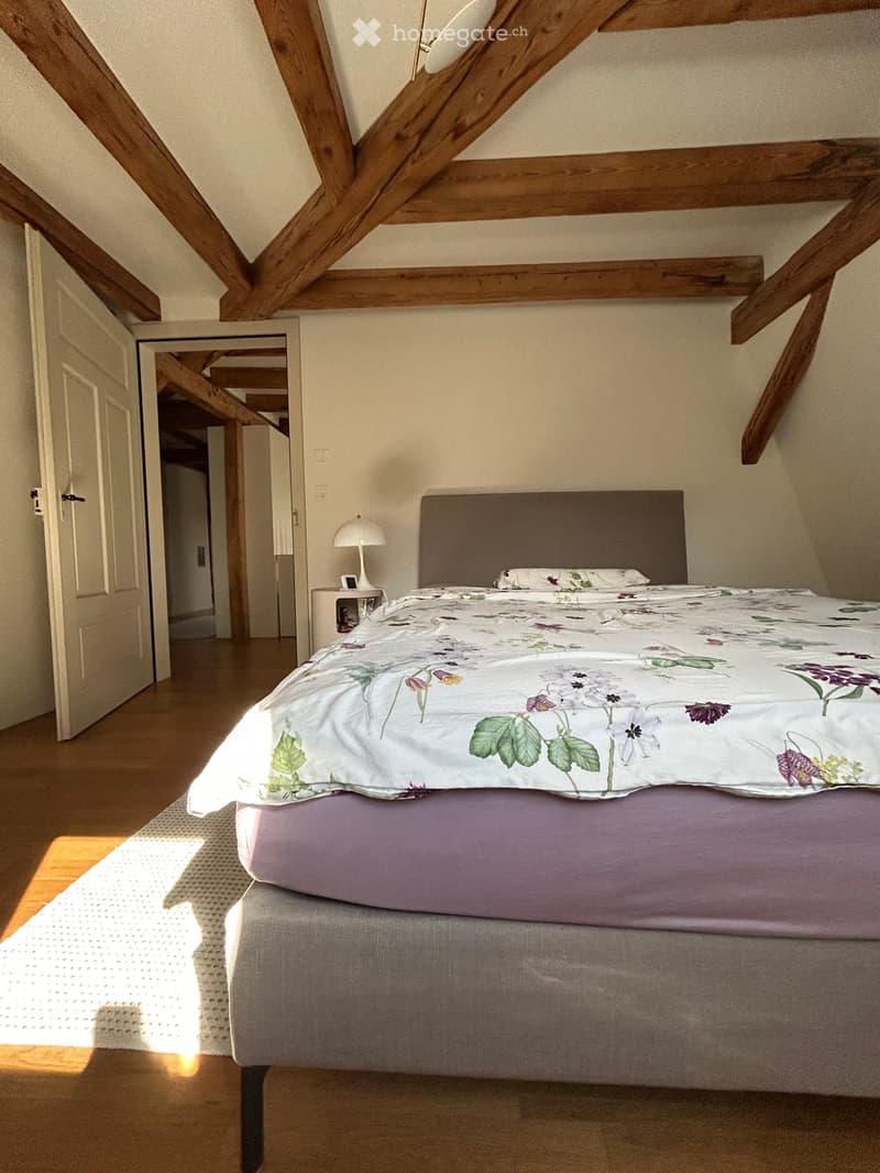 Cozy attic bedroom with wooden beams, a large bed with floral bedding, a nightstand with a lamp, and a small rug on the floor.