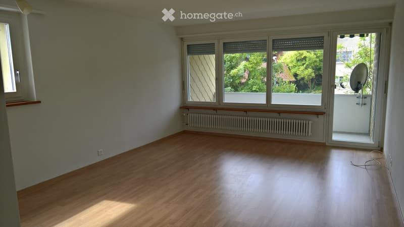 Empty room with wooden flooring, large windows, radiator, and a sliding glass door leading to a balcony.