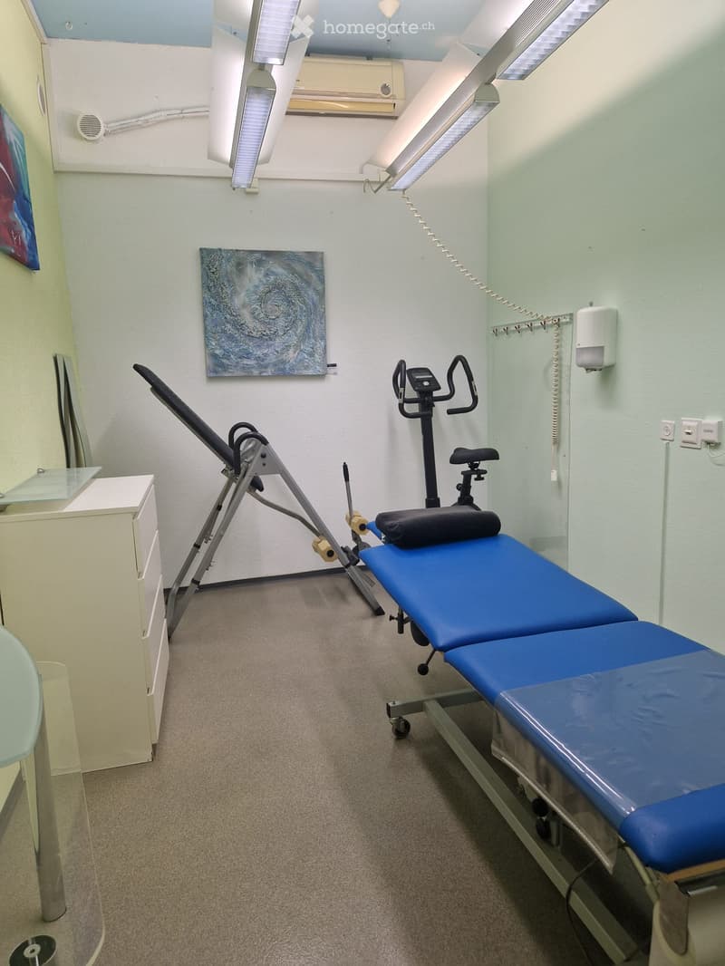 A clean, well-lit room with a treatment table, exercise machine, cabinet, mirror, and artwork on the walls.