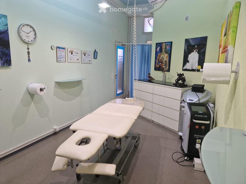 A well-lit medical or treatment room with a treatment table, a chair, a cabinet, a medical device labeled "Artmed", a mirror, a clock, a shelf, and several picture frames on the walls.