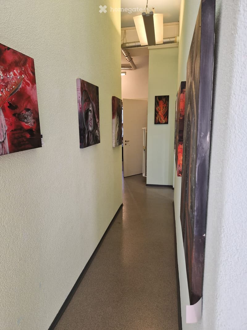 A narrow hallway with beige walls, carpeted floor, and several paintings displayed on both sides. A white door is visible at the end of the hallway.