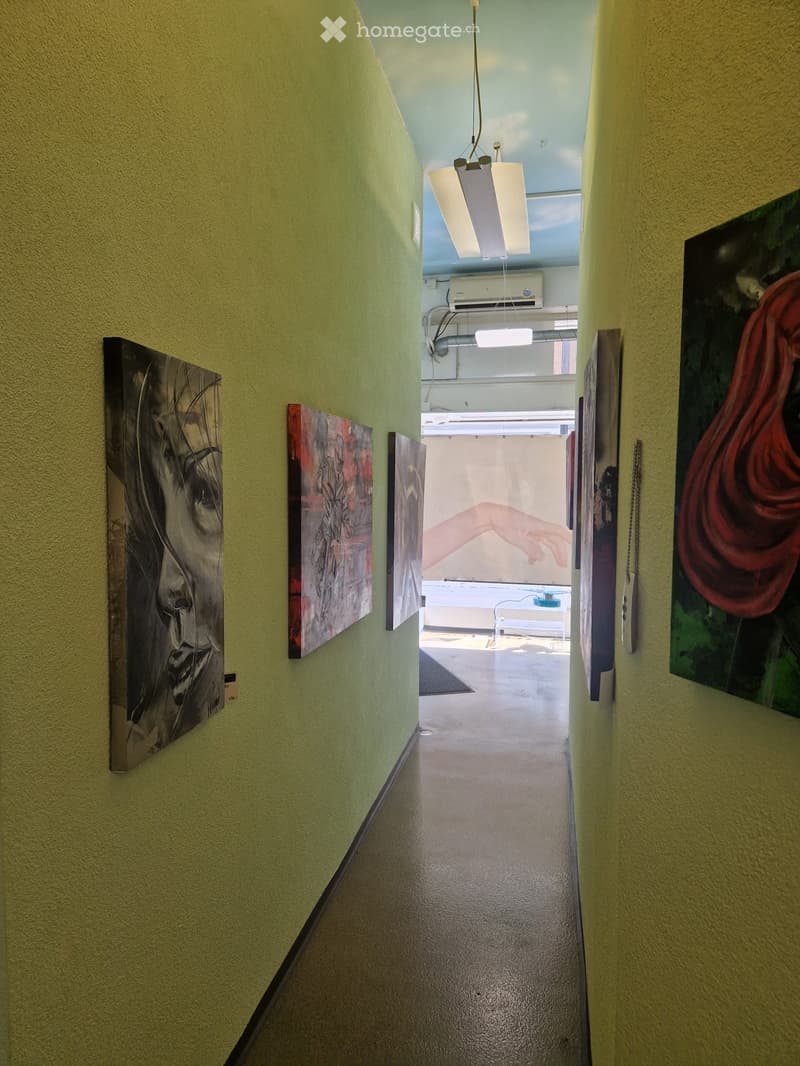 A narrow hallway with light green walls, several modern art pieces displayed on the walls, and a hanging light fixture. The hallway leads to an open area with more art and a visible table and chairs.