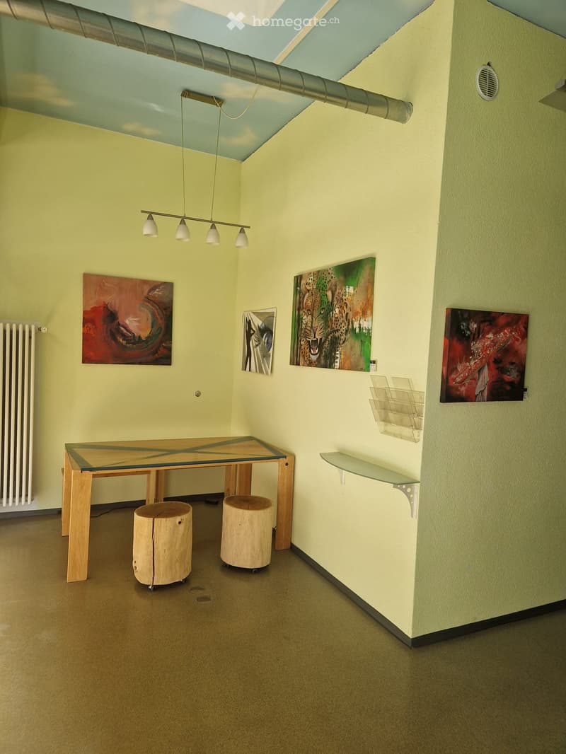 A modern, minimalist room with a glass-topped wooden table, two wooden stools, wall-mounted shelves, and several paintings on the walls.