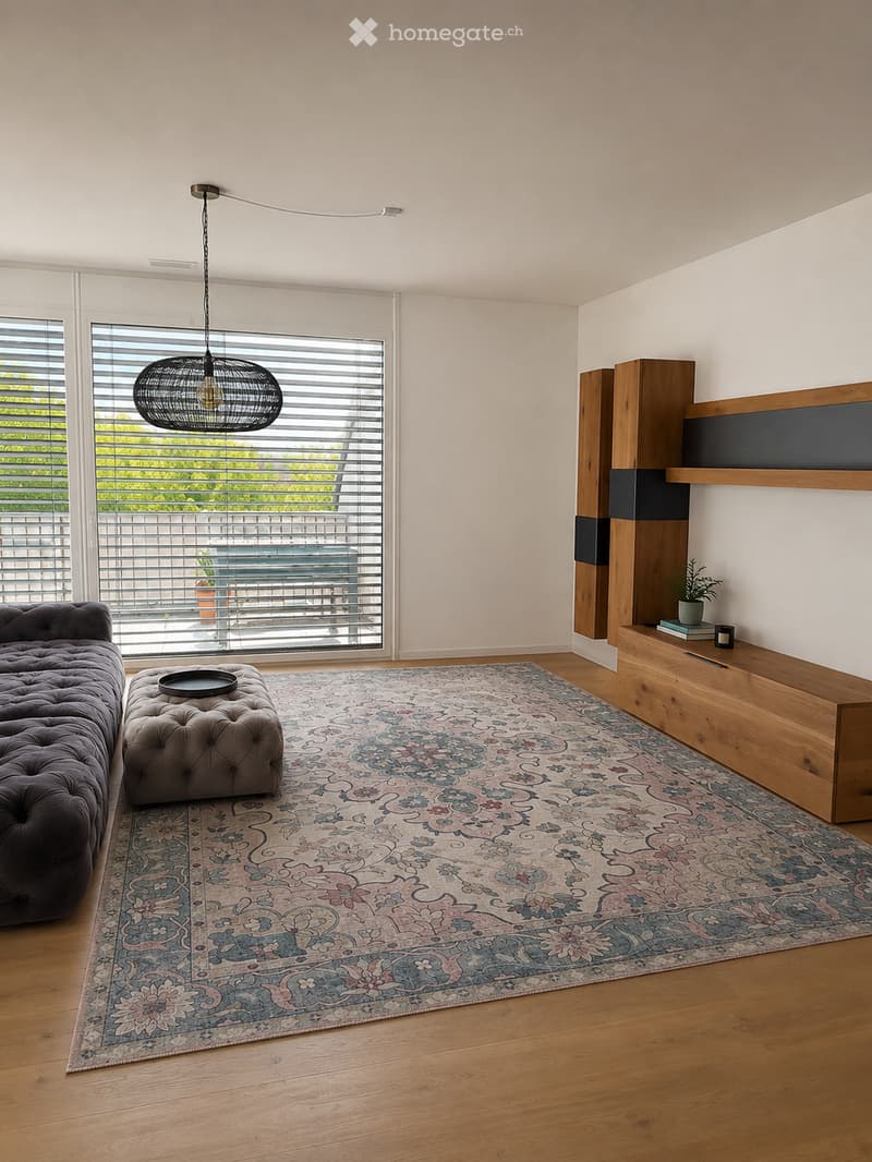 Modern living room with a large window, wooden floor, patterned rug, dark grey sofa, ottoman, wooden shelf, and pendant light.