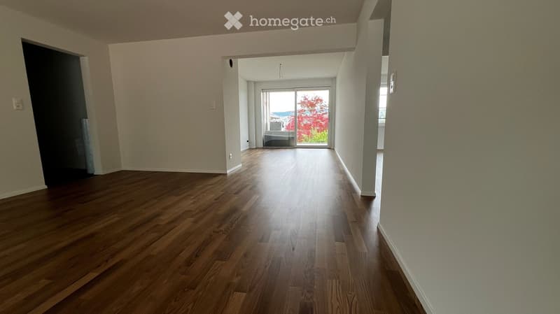 Empty hallway with wooden flooring, white walls, and a large window at the end leading to a view of trees and a body of water.