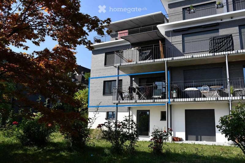 Modern two-story house with grey siding, multiple balconies, and a covered terrace. Features include a garage, outdoor furniture on the balconies, and surrounding greenery.