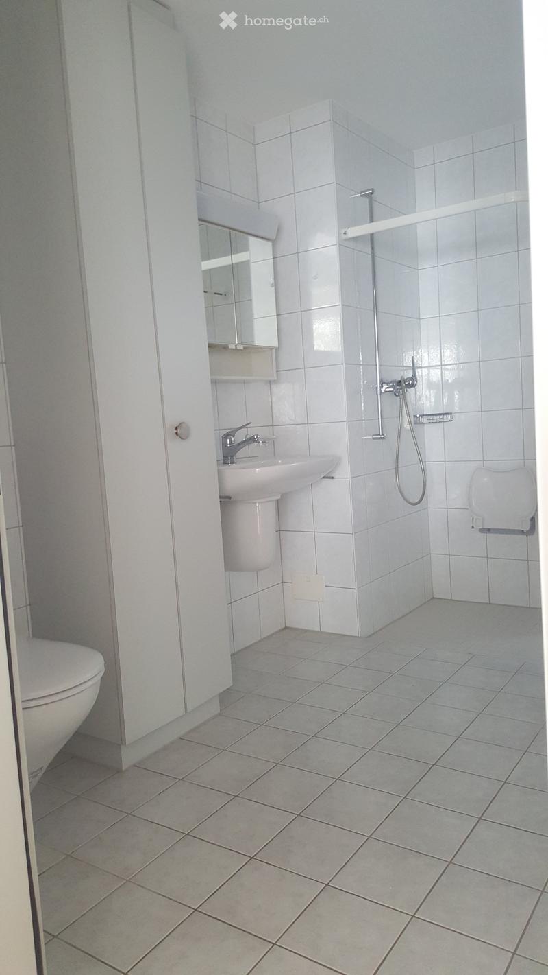 Modern bathroom with white tiled walls and floor, featuring a toilet, sink with mirror, and a shower area with a glass enclosure.