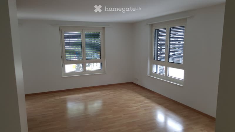 Empty room with wooden flooring and large windows with blinds, allowing natural light to enter.