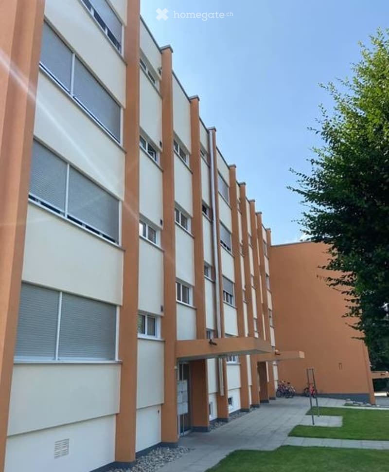 Modern multi-story residential building with beige and orange facade, large windows, and a covered walkway leading to the entrance.