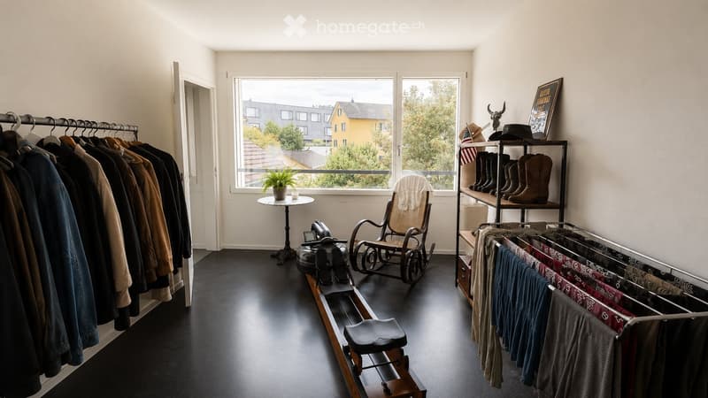 A well-lit room with a large window showing a view of the city, featuring a rocking chair, a small table with a plant, a clothing rack, a shelf with boots and hats, and a picture frame on the wall.