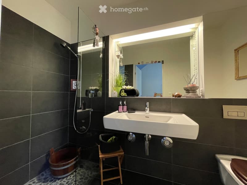 Modern bathroom with dark tiled walls, a double sink, a large mirror with built-in lighting, a shower area, and decorative items on the counter.