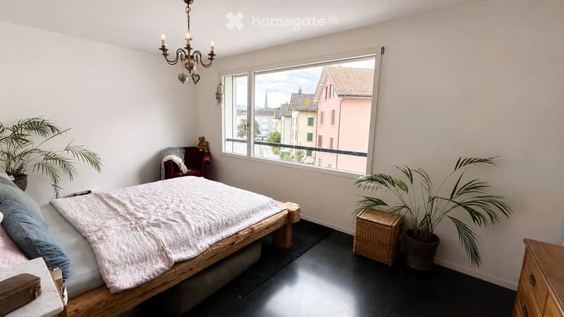 Bright bedroom with a wooden bed, a red armchair, a wicker basket, and large windows offering a city view.