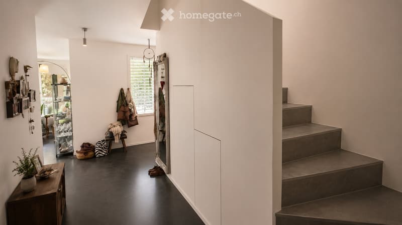 Modern hallway with dark flooring, white walls, staircase leading upwards, and various decorative items including a mirror, shoe rack, and small table with a plant.