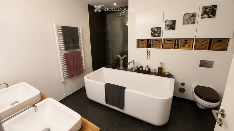 Modern bathroom with a large corner bathtub, a toilet, a sink, a towel rack with towels, a shower area, a mirror, wicker baskets on a shelf, framed pictures, a small plant, and a decorative statue.