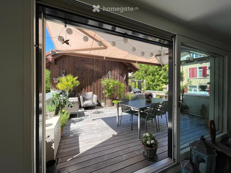 Balcony with wooden decking, outdoor furniture including a table, chairs, and a sofa, decorated with potted plants and flowers.