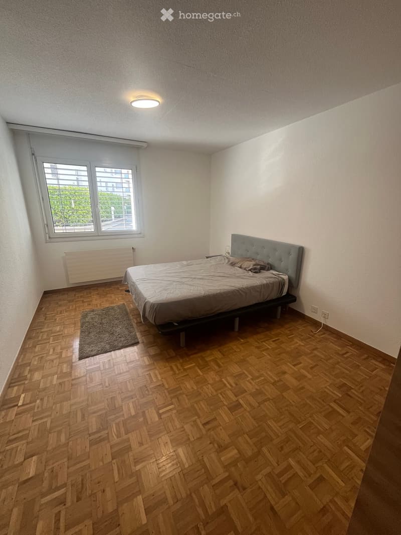Bedroom with wooden herringbone flooring, a single bed with a grey duvet and brown pillow, a grey headboard, a small brown rug, and a radiator under a window with blinds.