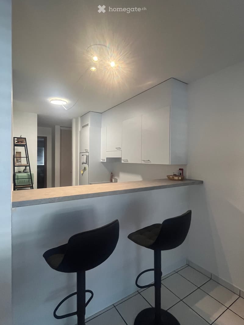 Modern kitchen with white cabinets, a countertop, and two black bar stools. A small shelf with items is visible in the background.