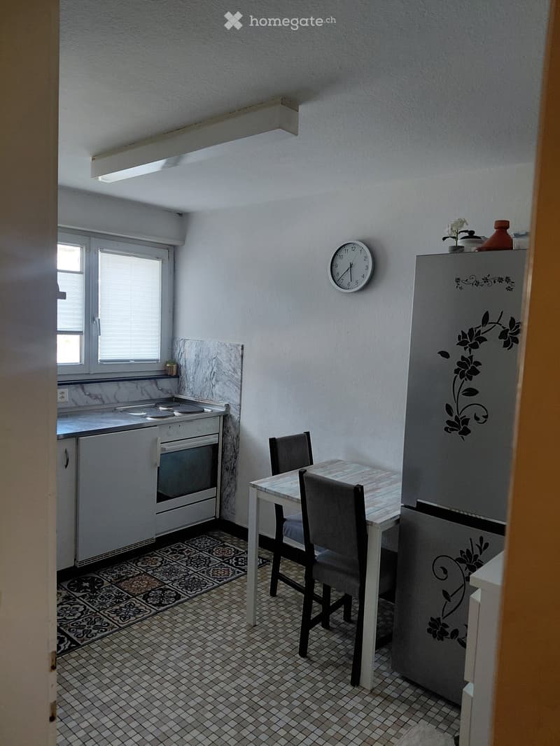 Compact kitchen with white cabinets, a small dining table with two chairs, a refrigerator, and an oven. The walls are white with a marble backsplash, and the floor is tiled.